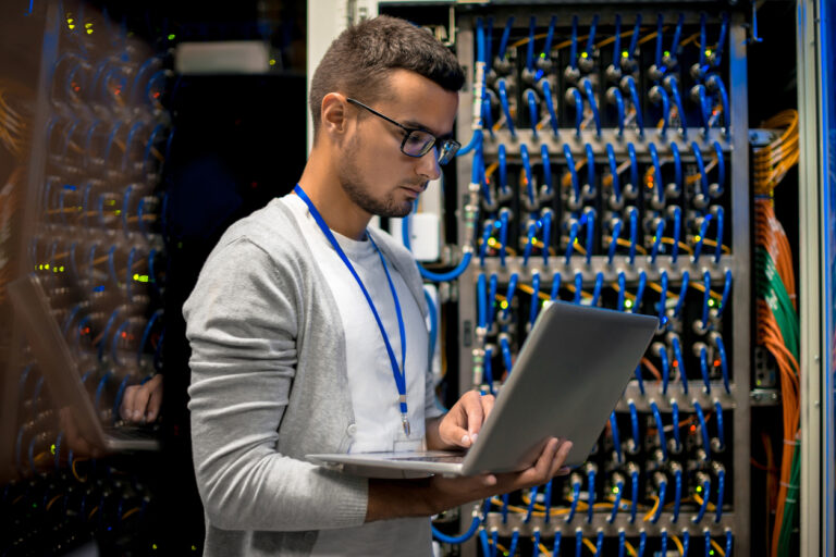 Side view  portrait of young man with laptop standing by server cabinet while working with supercomputer